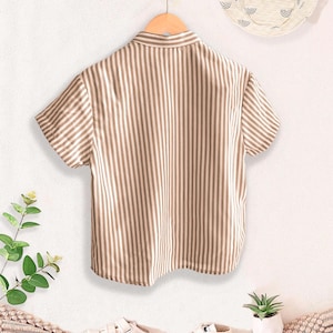 May include: A short-sleeved, button-down shirt with vertical brown and white stripes hangs on a wooden hanger. A matching sun hat with a white background and a pattern of curved lines is visible. White sneakers and a small potted plant are also in the image.