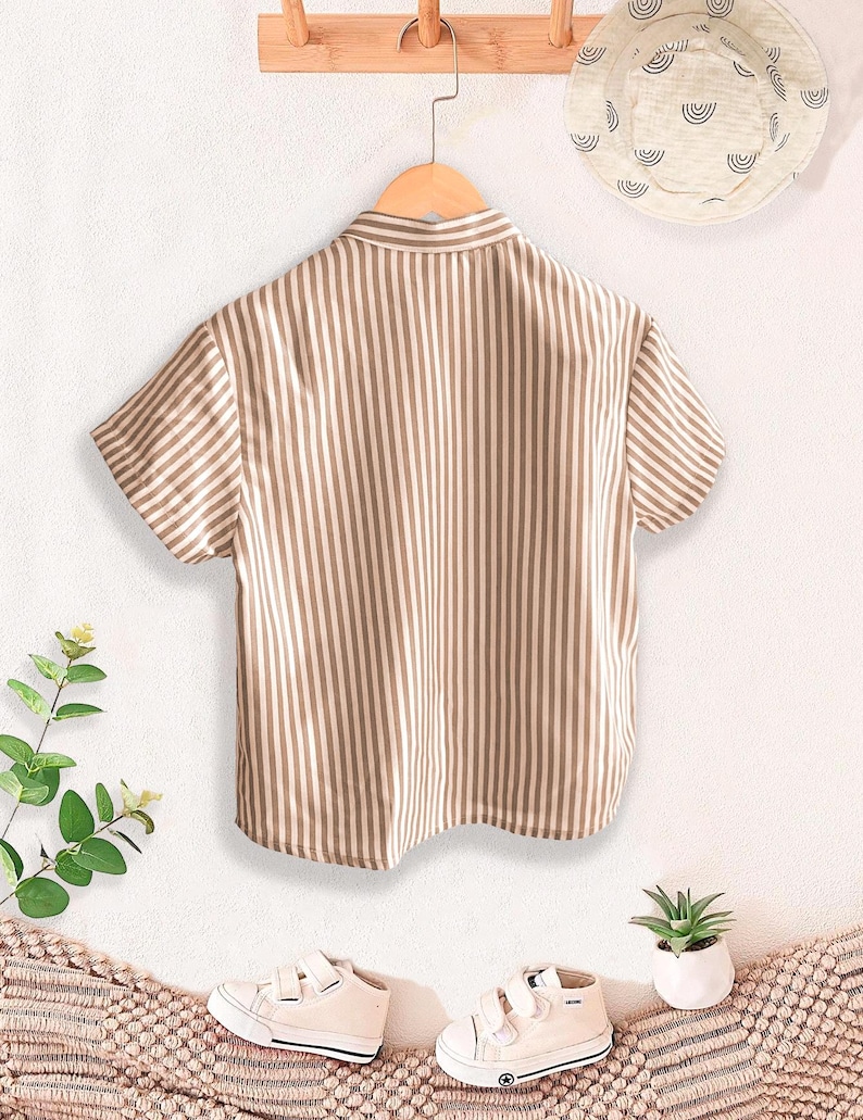 May include: A short-sleeved, button-down shirt with vertical brown and white stripes hangs on a wooden hanger. A matching sun hat with a white background and a pattern of curved lines is visible. White sneakers and a small potted plant are also in the image.