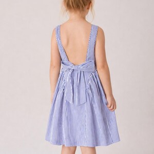 May include: A blue and white striped dress with a low back and a bow detail. The dress has a sleeveless design and a flared skirt. The child is facing away from the camera, with their hair in a bun.