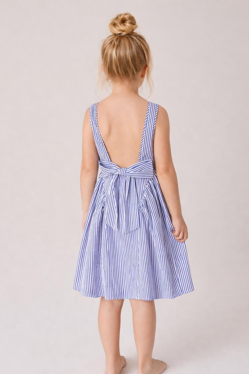 May include: A blue and white striped dress with a low back and a bow detail. The dress has a sleeveless design and a flared skirt. The child is facing away from the camera, with their hair in a bun.