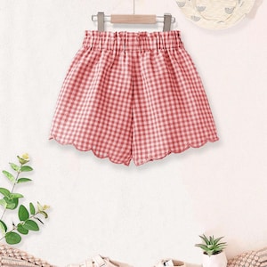 May include: Red and white gingham shorts with a scalloped hem, hanging on a wooden hanger. A matching sun hat with a rainbow design and white sneakers are also visible. The shorts are part of a children's clothing set.