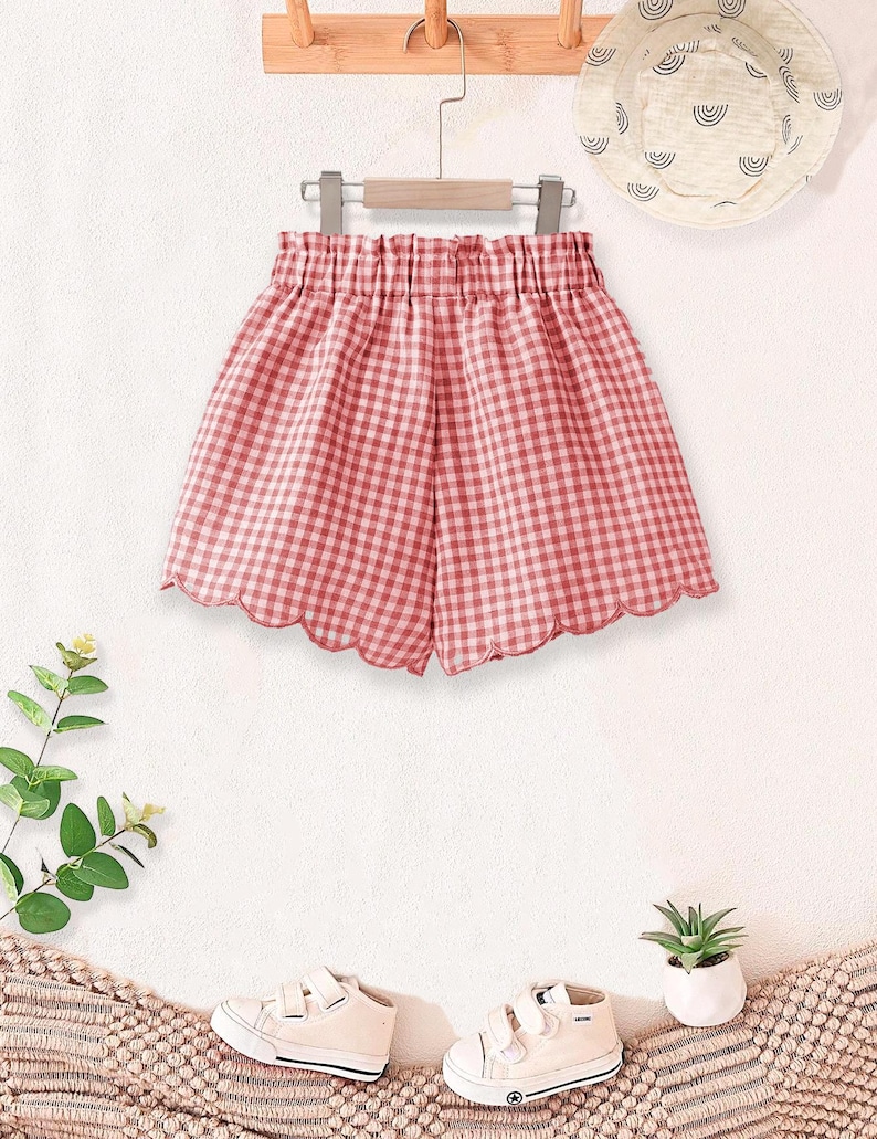 May include: Red and white gingham shorts with a scalloped hem, hanging on a wooden hanger. A matching sun hat with a rainbow design and white sneakers are also visible. The shorts are part of a children's clothing set.