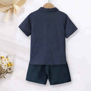 K&ouml;nnte beinhalten: Marineblaues kurz&auml;rmeliges Hemd mit Kragen und passendes Shorts-Set. Das Hemd hat ein strukturiertes, geripptes Design. Die Shorts sind in einem dunkleren Marineblau gehalten. Das Set h&auml;ngt an einem Holzkleiderb&uuml;gel.