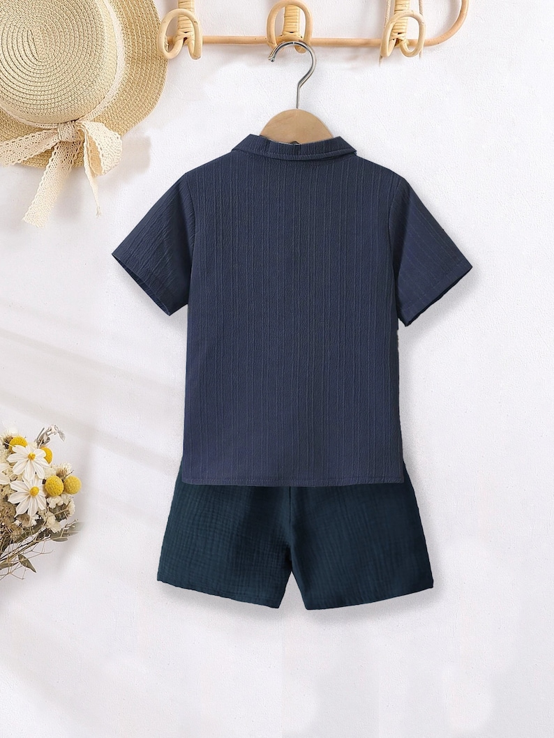 K&ouml;nnte beinhalten: Marineblaues kurz&auml;rmeliges Hemd mit Kragen und passendes Shorts-Set. Das Hemd hat ein strukturiertes, geripptes Design. Die Shorts sind in einem dunkleren Marineblau gehalten. Das Set h&auml;ngt an einem Holzkleiderb&uuml;gel.