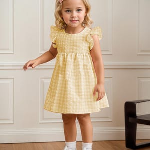 May include: A young child wearing a yellow gingham dress with ruffled sleeves and a gathered waist. The dress is paired with white socks and sneakers. The child has blonde hair and a pink hair clip.