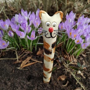 Könnte beinhalten: Eine Keramik-Katzenfigur mit einem weiß-braun gestreiften Körper, schwarzen Augen und einer roten Nase. Die Figur steht in einem Beet mit violetten Krokussen.