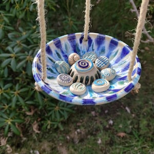 Può includere: Una mangiatoia per uccelli in ceramica sospesa con una ciotola a motivi blu e bianchi. La ciotola è riempita di piccole pietre decorative. La mangiatoia è sospesa da una corda naturale, creando un'estetica rustica. Circondata da fogliame verde.