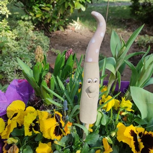 Könnte beinhalten: Ein Keramik-Gartenzwerg mit einem rosa-weißen gepunkteten Hut und einem lächelnden Gesicht. Der Zwerg ist umgeben von gelben Stiefmütterchen und grünen Pflanzen in einem violetten Topf.