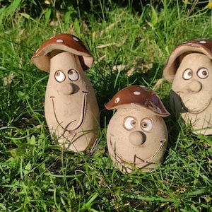 Könnte beinhalten: Drei Keramik-Pilzfiguren mit braunen und weißen Details. Jeder Pilz hat ein lächelndes Gesicht mit großen Augen und einer weißen Nase. Die Pilze sitzen auf einem Bett aus grünem Gras.