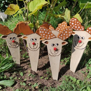 Könnte beinhalten: Vier Gartenstecker aus Keramik in Form von Rentieren. Jeder Stecker zeigt ein lächelndes Rentiergesicht mit roter Nase, braunem Geweih und cremefarbenem Körper. Zwei der Rentiere haben weiße Punkte auf ihrem Geweih. Die Stecker stehen in einer Grasfläche.