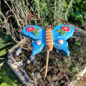Könnte beinhalten: Ein blauer Keramik-Schmetterlings-Gartenpfahl mit roten Blumen und rosa Akzenten. Der Schmetterling hat einen braunen Streifen in der Mitte seines Körpers.