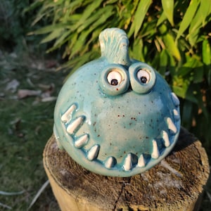 Könnte beinhalten: Eine tealblaue Keramikmonsterfigur mit weißen Zähnen und großen weißen Augen. Das Monster sitzt auf einem Baumstumpf.