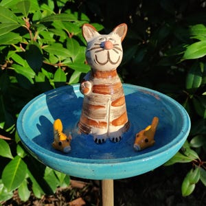 Könnte beinhalten: Ein Vogelbad aus Keramik mit einer blauen Schale und einer Katzenfigur. Die Katze ist braun und weiß mit einem gestreiften Muster und sitzt am Rand der Schale. Zwei kleine gelbe Fischfiguren befinden sich in der Schale.