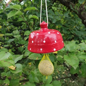Könnte beinhalten: Roter Keramik-Vogel futterspender mit weißen Punkten, der an einem Baumzweig hängt. Ein gelber Talgkuchen befindet sich im Futterspender.