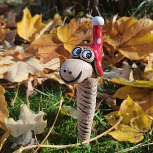 Può includere: Una statuetta di verme in ceramica con un cappello a pois rossi e bianchi. Il verme ha un viso sorridente con occhi grandi e si trova in un letto di foglie autunnali.