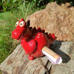Könnte beinhalten: Eine rote Keramik-Drachenfigur mit weißen Punkten und einem breiten Lächeln. Der Drache hat ein Loch an der Seite, und ein zusammengerolltes Stück Papier schaut heraus.