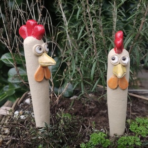Könnte beinhalten: Zwei skurrile Gartenstecker in Form von Hähnen aus Keramik. Jeder Stecker hat einen beigefarbenen Körper, einen roten Kamm, einen gelben Schnabel und orangefarbene Kehllappen. Die Stecker sind in einem Gartenbeet mit grünem Laub im Hintergrund gepflanzt.