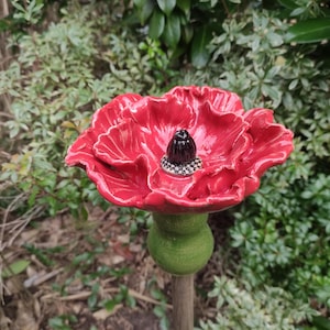 Può includere: Un fiore di papavero in ceramica rossa con un centro nero. Il fiore è su uno stelo verde ed è attaccato a un paletto di legno.