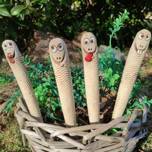 Può includere: Quattro figurine di serpente in ceramica con volti dipinti e un'espressione fantasiosa. I serpenti sono in un cesto intrecciato e hanno una superficie testurizzata.