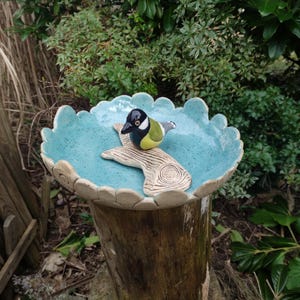 Könnte beinhalten: Ein Vogelbad aus Keramik mit blauer Glasur und weißem Rand. Ein kleiner Vogel aus Keramik sitzt auf einem brettartigen Sitz in der Mitte des Bades.
