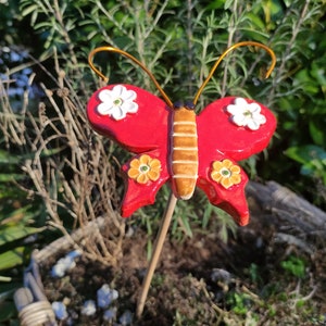 Könnte beinhalten: Roter Keramik-Schmetterlings-Gartenpfahl mit weißen und gelben Blüten. Der Schmetterling hat braune Streifen auf seinen Flügeln.