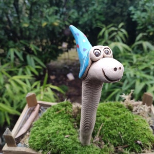 Könnte beinhalten: Ein skurriler Keramik-Gartenzwerg mit einem blau-weiß gepunkteten Hut, der mit großen Augen lächelt. Der Zwerg steht in einem Bett aus grünem Moos.