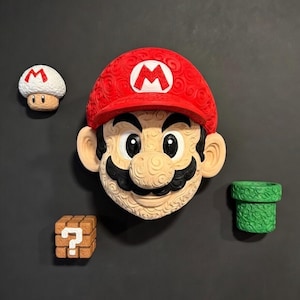 Puede incluir: Un conjunto de arte mural temático de Super Mario Bros. Incluye una gran cabeza de Mario con gorra roja, una seta, un bloque de interrogación y una tubería verde. Las piezas tienen textura y parecen ser de material duradero.