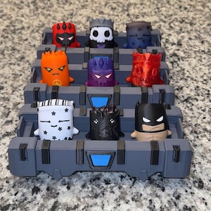Peut inclure: Un plateau de rangement en plastique gris avec des accents bleus contenant neuf petites figurines de personnages stylisées. Les figurines présentent divers motifs, dont un crâne rouge, un fantôme blanc étoilé, une créature noire et un masque.