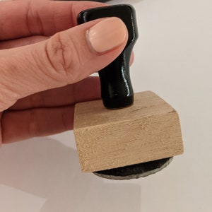 May include: A wooden stamp with a black handle. The stamp has a rectangular base and a round ink pad.