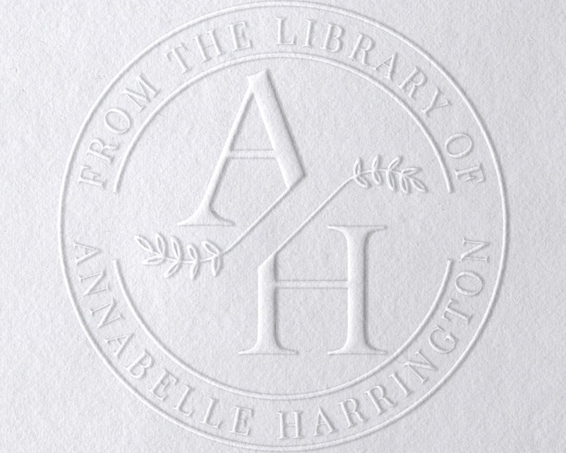 Puede incluir: Sello circular blanco en relieve con el texto "From the Library of Annabelle Harrington" y las iniciales "AH" con una peque&ntilde;a rama de hojas entre ellas.