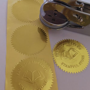 May include: Gold foil sticker seals with a book design and the text "From the Library of Stampful Store". The stickers are on a white surface next to a silver metal stamp.
