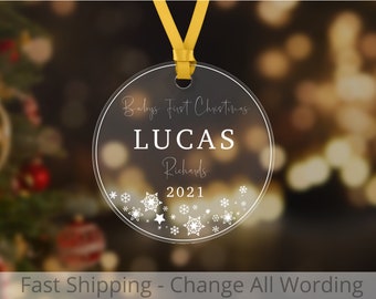 May include: Clear round Christmas ornament with a yellow ribbon. The ornament has white snowflakes and a star design. The text "Baby's First Christmas LUCAS Richards 2021" is printed on the ornament.