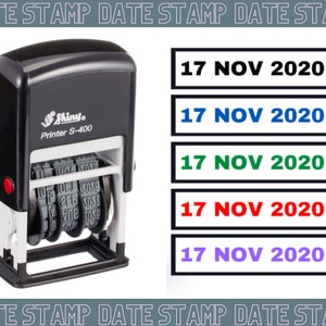 Op de afbeelding: Een zwarte datumstempel met een Shiny Printer S-400 label. De datumstempel heeft een rode knop en drie draaiende wielen met cijfers. De datumstempel is ingesteld op 17 november 2020. De afbeelding toont ook vijf verschillende gekleurde rechthoeken met de datum "17 NOV 2020" in zwart gedrukt.
