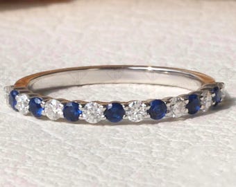 May include: A silver ring with alternating round blue and clear stones. The ring is set against a white background with a blurred background of wheat stalks.