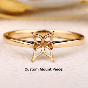 Puede incluir: Un anillo dorado con un diseño calado distintivo en el centro, con una pieza de montaje personalizada. La banda del anillo es delgada, y el diseño central tiene forma de flor estilizada. El texto "Custom Mount Piece!" es visible.