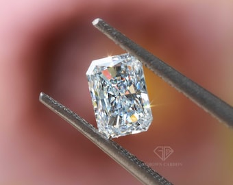 May include: A clear, rectangular, emerald-cut diamond held by tweezers against a blurred pink and green background. The diamond is reflecting light and has a brilliant sparkle. The text "CROWN CARBON" is visible in the lower right corner of the image.
