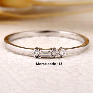Könnte beinhalten: Ein zarter Silberring mit drei kleinen Diamanten. Der mittlere Diamant ist rechteckig, flankiert von zwei runden Diamanten. Der Text "Morse code - L!" befindet sich darunter.