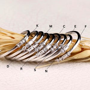 Könnte beinhalten: Eine Sammlung silberfarbener, gebogener Armreifen, die jeweils mit klaren, funkelnden Edelsteinen verziert sind. Die Armreifen sind in einer Reihe angeordnet und präsentieren ihr elegantes Design und die unterschiedlichen Edelsteinanordnungen. Das Grown Carbon-Logo ist unten rechts sichtbar.