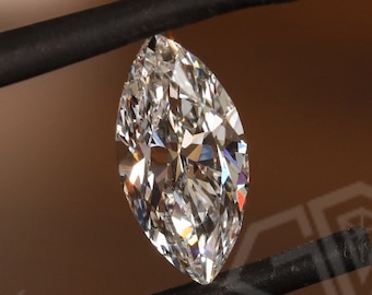Könnte beinhalten: Ein klarer, marquiseförmiger Diamant, der mit einer Pinzette gehalten wird. Der Diamant ist funkelnd und hat einen Brillantschliff. Der Hintergrund ist eine braune Oberfläche mit dem aufgedruckten Text "GROWN CARBON".