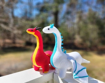 May include: Two plush dragon toys. One is red with yellow accents, the other is white with blue accents. Both have long necks, small wings, and are standing on a white surface. The background is blurred with green and blue tones.
