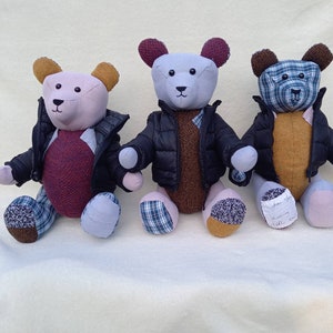 May include: Three handmade teddy bears wearing black puffer jackets with plaid accents. The bears are sitting on a white surface. The bear on the left is pink with a burgundy sweater, the middle bear is light purple with a brown sweater, and the bear on the right is brown with a yellow sweater.
