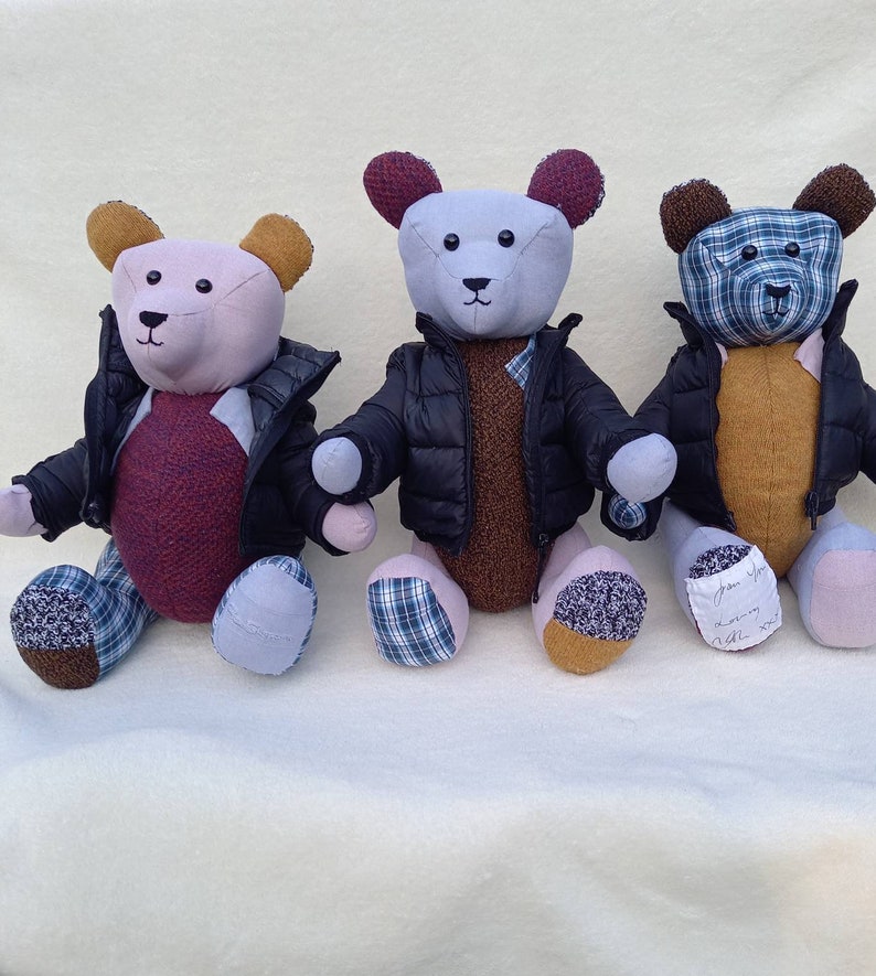 May include: Three handmade teddy bears wearing black puffer jackets with plaid accents. The bears are sitting on a white surface. The bear on the left is pink with a burgundy sweater, the middle bear is light purple with a brown sweater, and the bear on the right is brown with a yellow sweater.