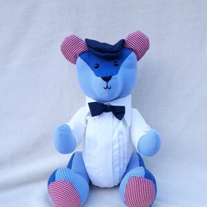 May include: A blue and white patchwork teddy bear wearing a white shirt, a black bow tie, and a black hat. The bear has red and white checkered ears and feet.