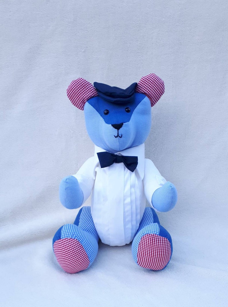 May include: A blue and white patchwork teddy bear wearing a white shirt, a black bow tie, and a black hat. The bear has red and white checkered ears and feet.
