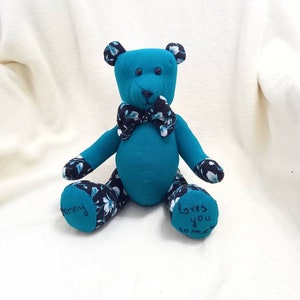 May include: A teal blue teddy bear with a black and white floral pattern on its paws. The bear is wearing a black and white bow tie and has the words "Loves you so much" embroidered on one paw and "Mommy" embroidered on the other paw.