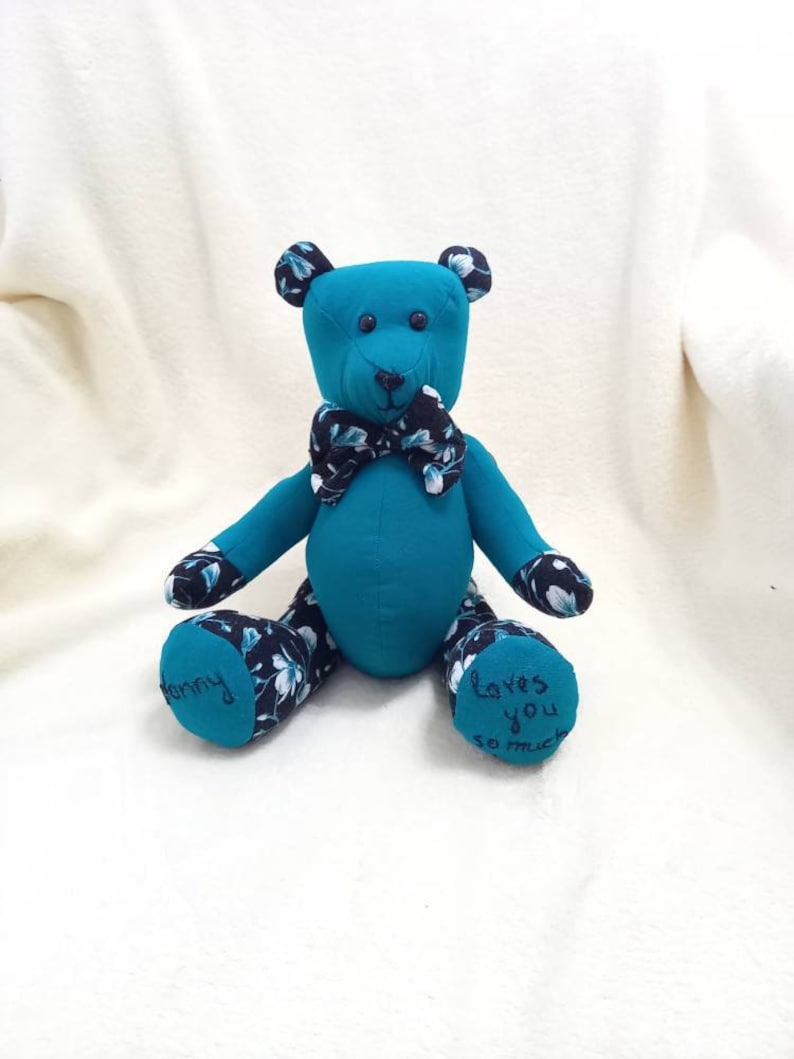 May include: A teal blue teddy bear with a black and white floral pattern on its paws. The bear is wearing a black and white bow tie and has the words "Loves you so much" embroidered on one paw and "Mommy" embroidered on the other paw.