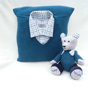 May include: A teal blue knitted sweater cushion with a white and brown checked shirt inside. A teal blue teddy bear with a white face and blue embroidered feet sits in front of the cushion. The bear's feet are embroidered with "Granddad" and "1936-2024".