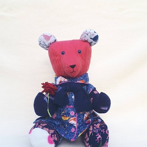May include: A pink knitted teddy bear wearing a blue and floral dress with a navy blue cardigan and a bow. The bear has white fabric ears with floral patterns and is holding a red flower.