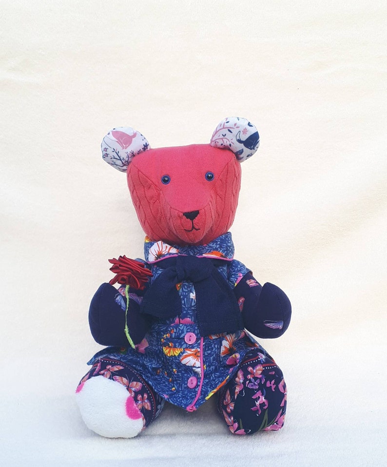 May include: A pink knitted teddy bear wearing a blue and floral dress with a navy blue cardigan and a bow. The bear has white fabric ears with floral patterns and is holding a red flower.