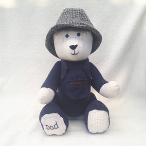 May include: A blue and white teddy bear wearing a tweed hat and a blue shirt with a pocket. The bear's foot is embroidered with the word "Dad".
